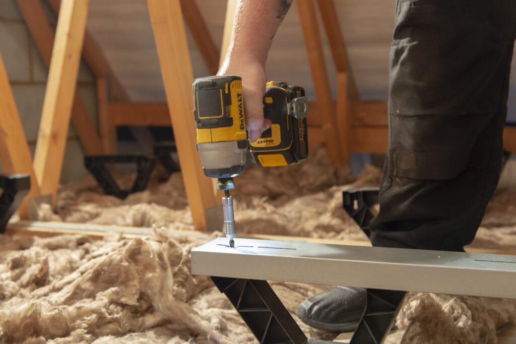 A person drilling a loft floor raiser into the floor of their loft with open insulation in the background.