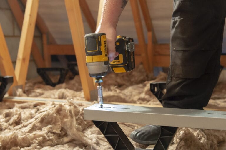 A person drilling a loft floor raiser into the floor of their loft with open insulation in the background.